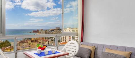 A balcony with a view of the sea and cityscape, a table with a vase of flowers, and a sofa with cushions.