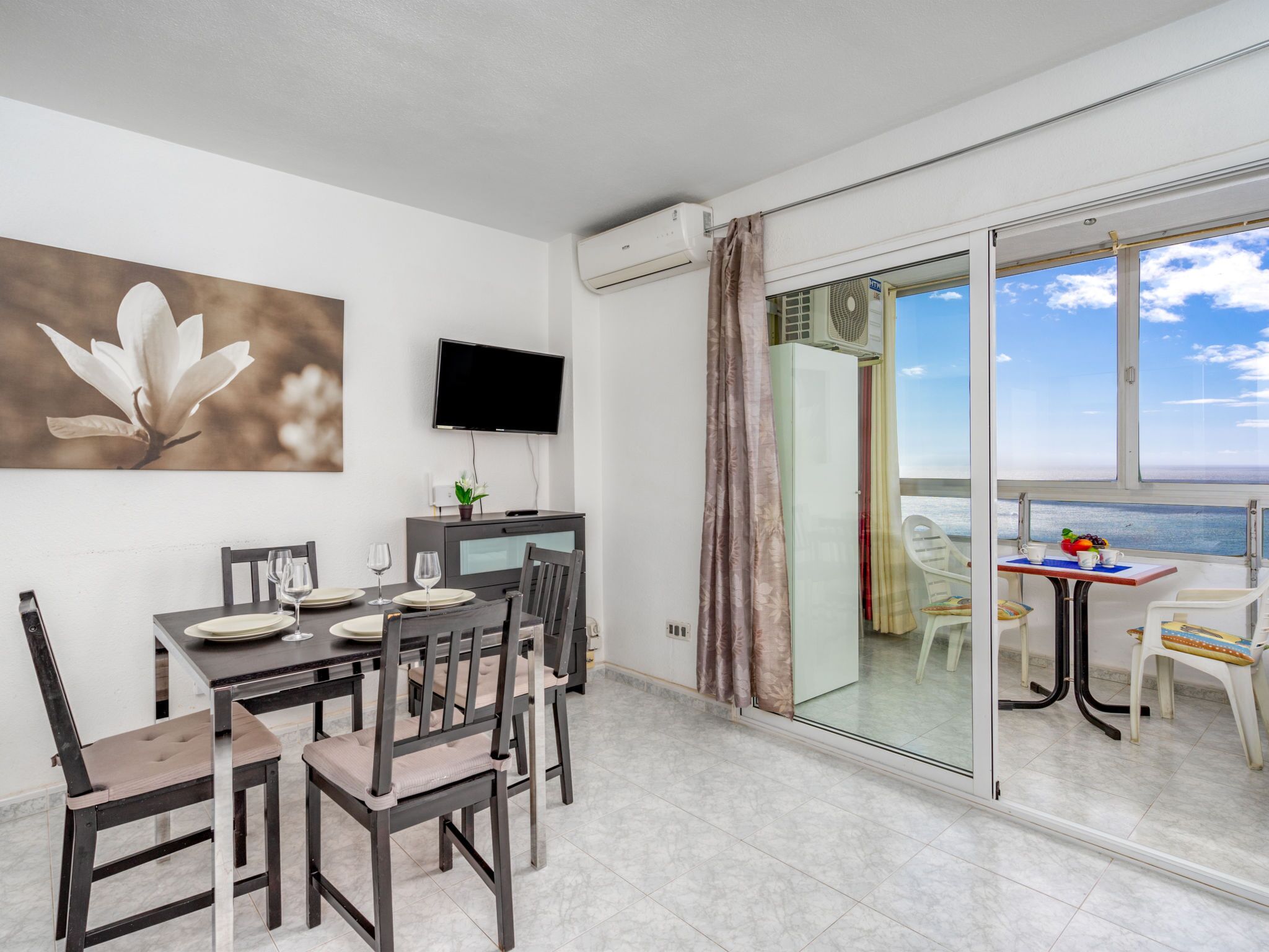 A dining area with a table set for four, a flat-screen TV mounted on the wall, and a sliding glass door leading to a balcony with a view of the sea.