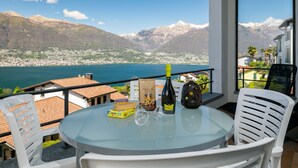 A balcony with a glass table, white chairs, a bottle, and a box of chocolates. - Residenza Miralago Apt. B16 by Interhome (Piazzogna)