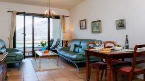 A modern living room with a dining area, green leather sofas, a chandelier, and a scenic mountain view through the glass door. - Residenza Miralago Apt. B16 by Interhome (Piazzogna)