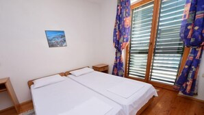 A bedroom with two single beds, wooden flooring, a small bedside table, a nightstand, a window with blinds, and a colorful curtain. - Apartment Orebić by Interhome (Orebić)
