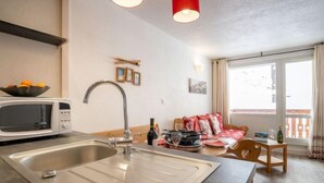 Cuina moderna amb microones, rentamicroplates i llums penjants vermelles. Hi ha una zona de menjador amb taula i cadires, i un sofà.