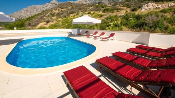 Une piscine bordée de chaises longues rouges et agrémentée d’un parasol blanc, le tout entouré de collines rocheuses.