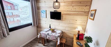 Ein gemütliches Zimmer mit einer Holzwand, einem Fernseher über einem Tisch für zwei Personen, einem Fenster mit Vorhängen und einer kleinen Pflanze auf einem Nachttisch.