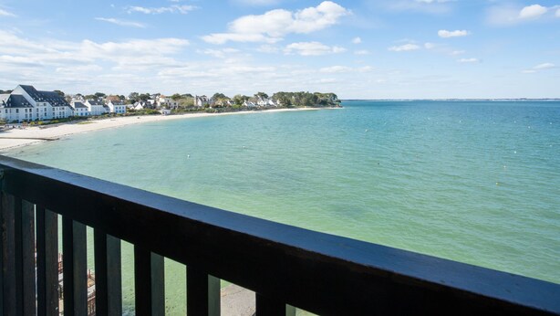 A coastal view with a wooden deck railing, a sandy beach, and a row of white buildings along the shore. - Bellevue by Interhome (Quiberon)