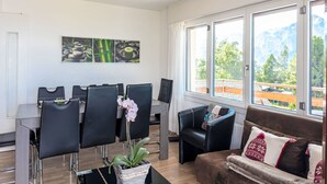 Ein modernes Wohnzimmer mit Essbereich, großen Fenstern und Bergblick sowie einer Deckenleuchte.