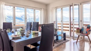 Ein Essbereich mit einem Tisch für vier Personen, großen Fenstern mit Bergblick und einem Balkon mit Holzgeländer.