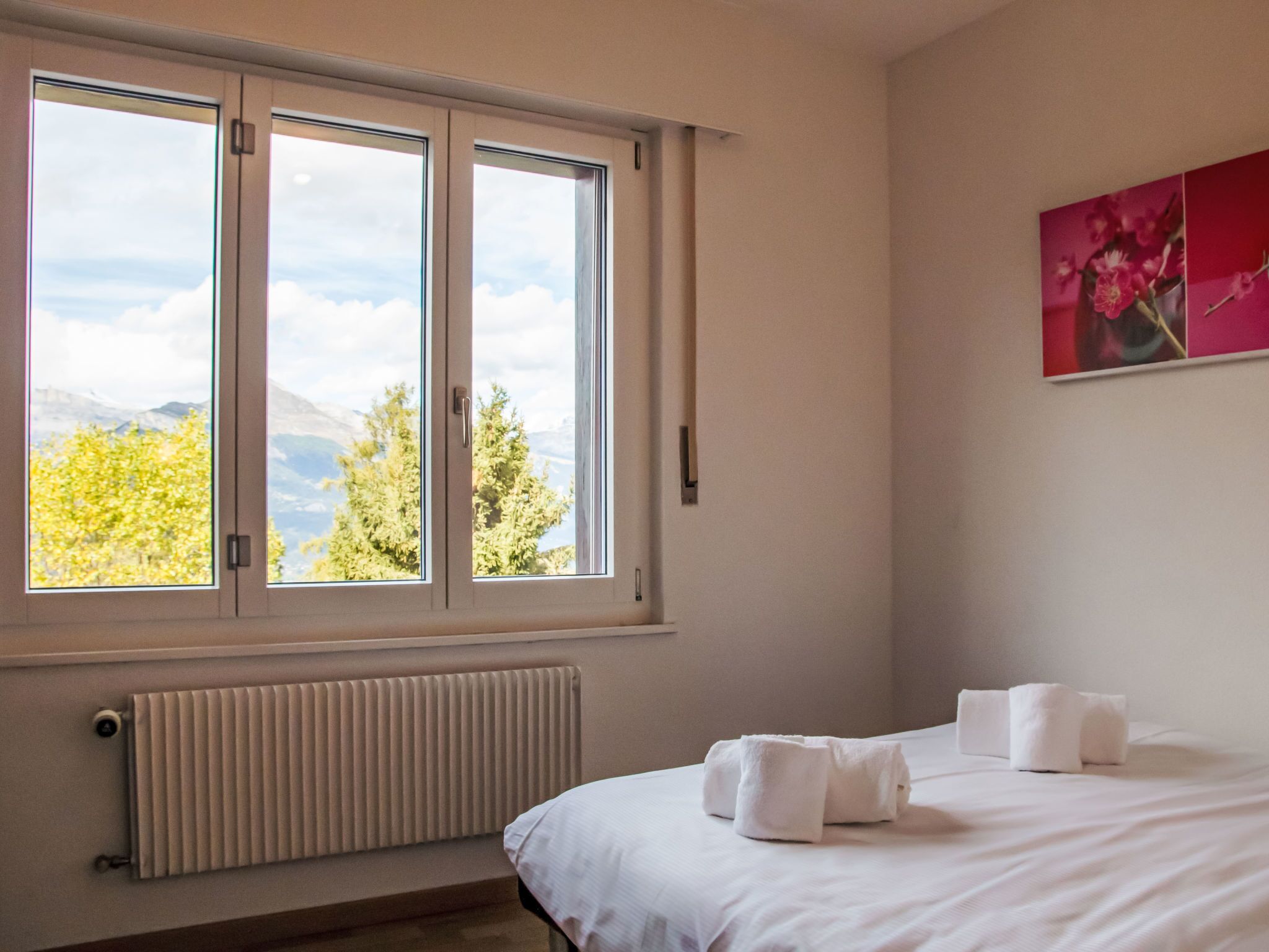 A hotel room with a large window, a radiator, a bed with white linens, and a painting on the wall.