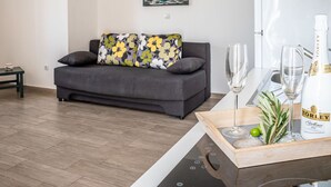 Un salon moderne avec un canapé gris, un plaid à motifs floraux, un tableau représentant un paysage côtier et un coin bar avec une bouteille de vin et des verres.