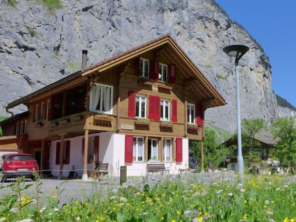 Eine zweistöckige Holzhütte mit roten Fensterläden, einem Balkon und einem Berg im Hintergrund.