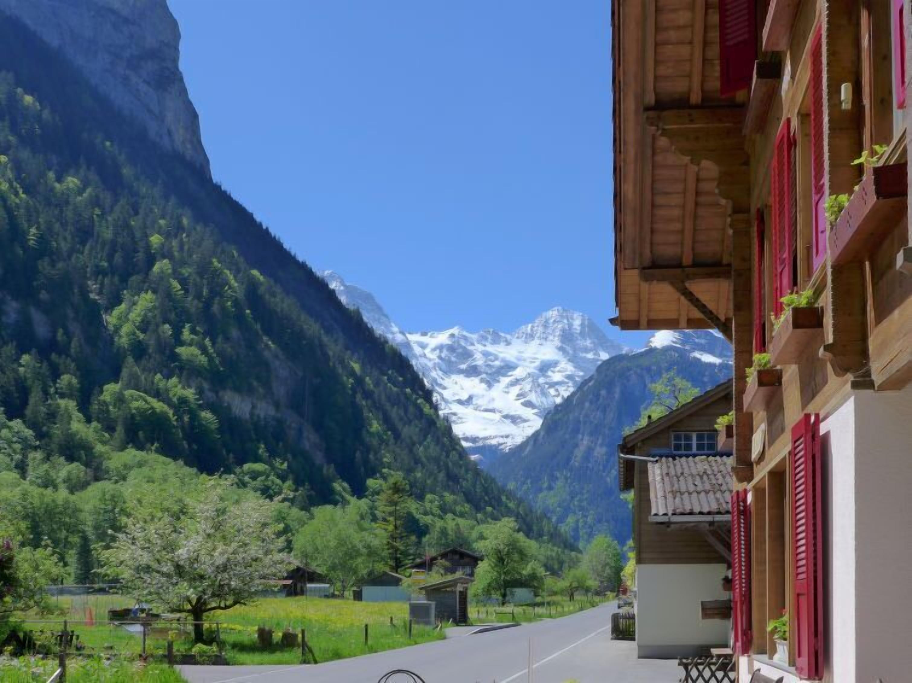 A mountain village with traditional wooden houses, a clear road, and snow-capped peaks in the background.