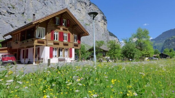 A chalet-style building with a wooden facade, multiple windows, and a balcony. It is situated in a mountainous area with a clear sky and a field of wildflowers in the foreground. - Apartment Rösli by Interhome (Lauterbrunnen)