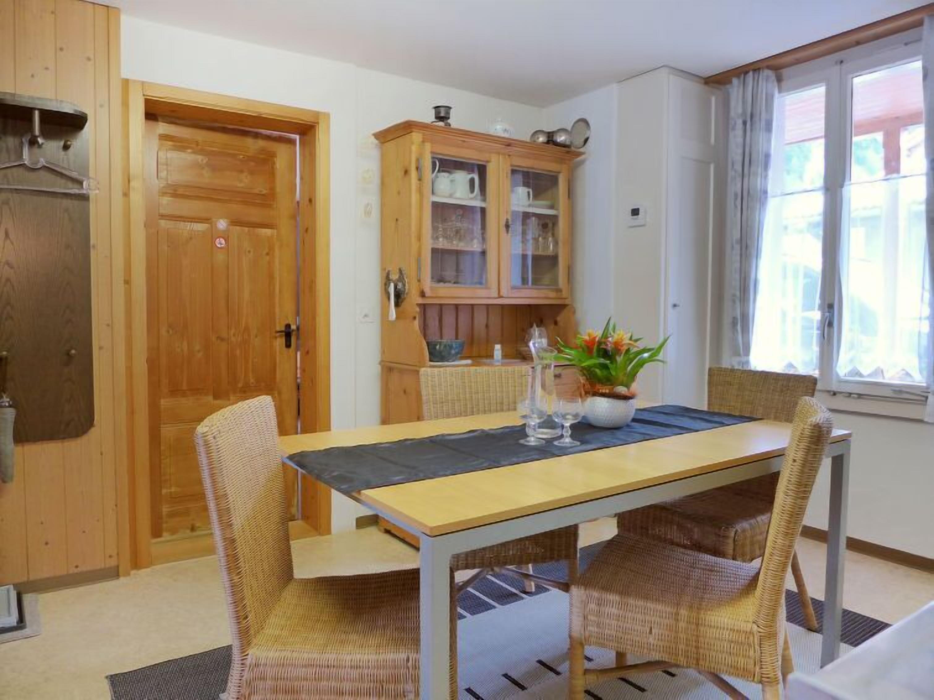 Un comedor con mesa de madera y sillas de mimbre, un armario con puertas de cristal y una ventana con cortinas.