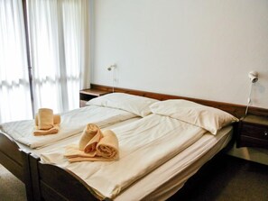 A neatly made bed with two pillows and two folded towels, a wooden headboard, a bedside table, and a power outlet on the wall.