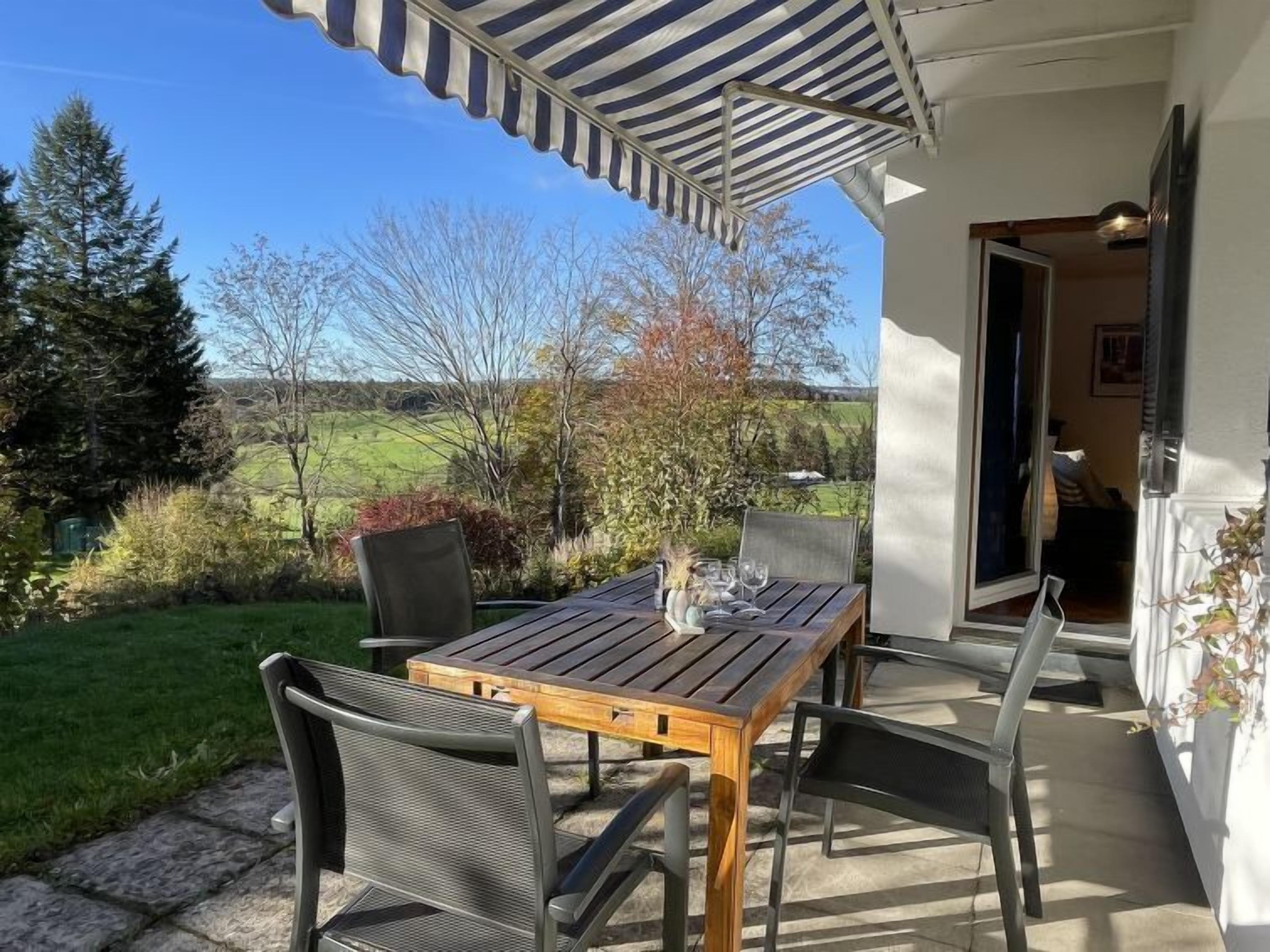 En udendørs terrasse med et træbord og stole, en stribet markise, og udsigt over et grønt landskab.