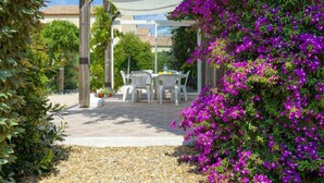Un patio al aire libre con una mesa y sillas, rodeado de exuberante follaje y flores moradas.