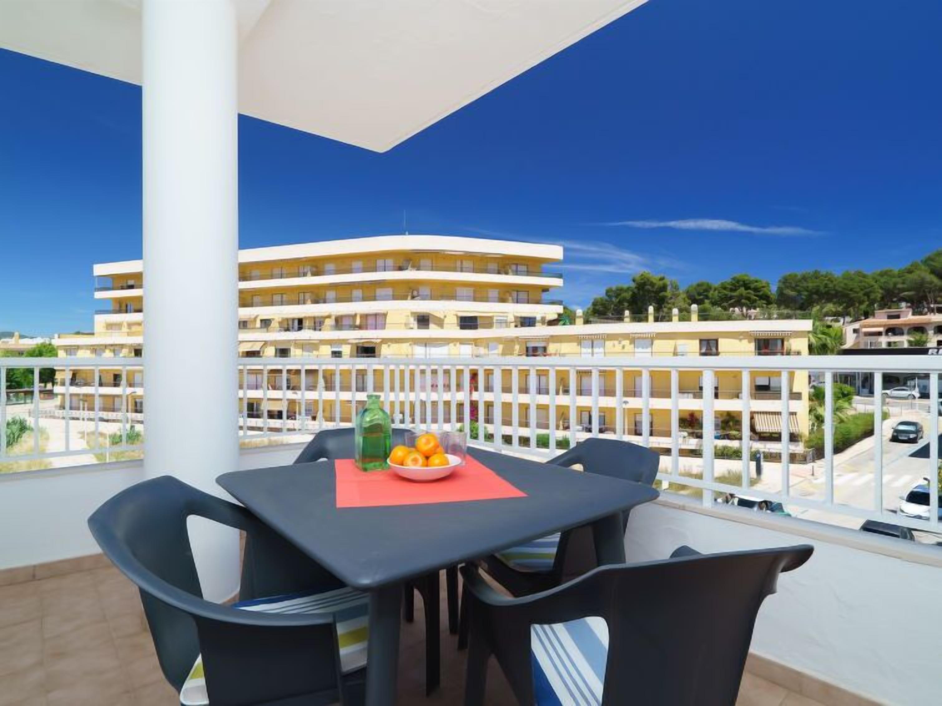 A balcony with a table and chairs, a bowl of fruit, and a bottle. The balcony overlooks a building with multiple floors and balconies.