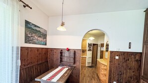 Une pièce comprenant un banc en bois, une table recouverte d’un chemin de table à motifs rouges et blancs, un lit avec une literie rose et une armoire en bois.