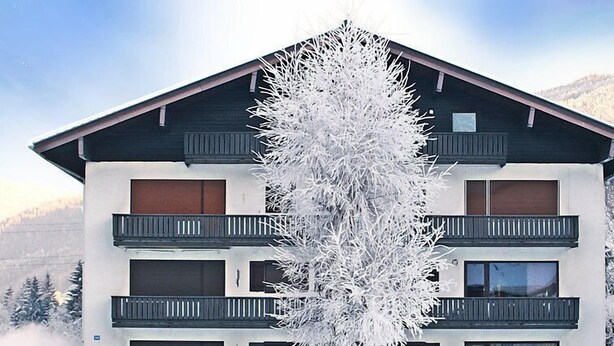 Un immeuble à plusieurs étages doté de balcons, avec un arbre devant et une chaîne de montagnes en arrière-plan.