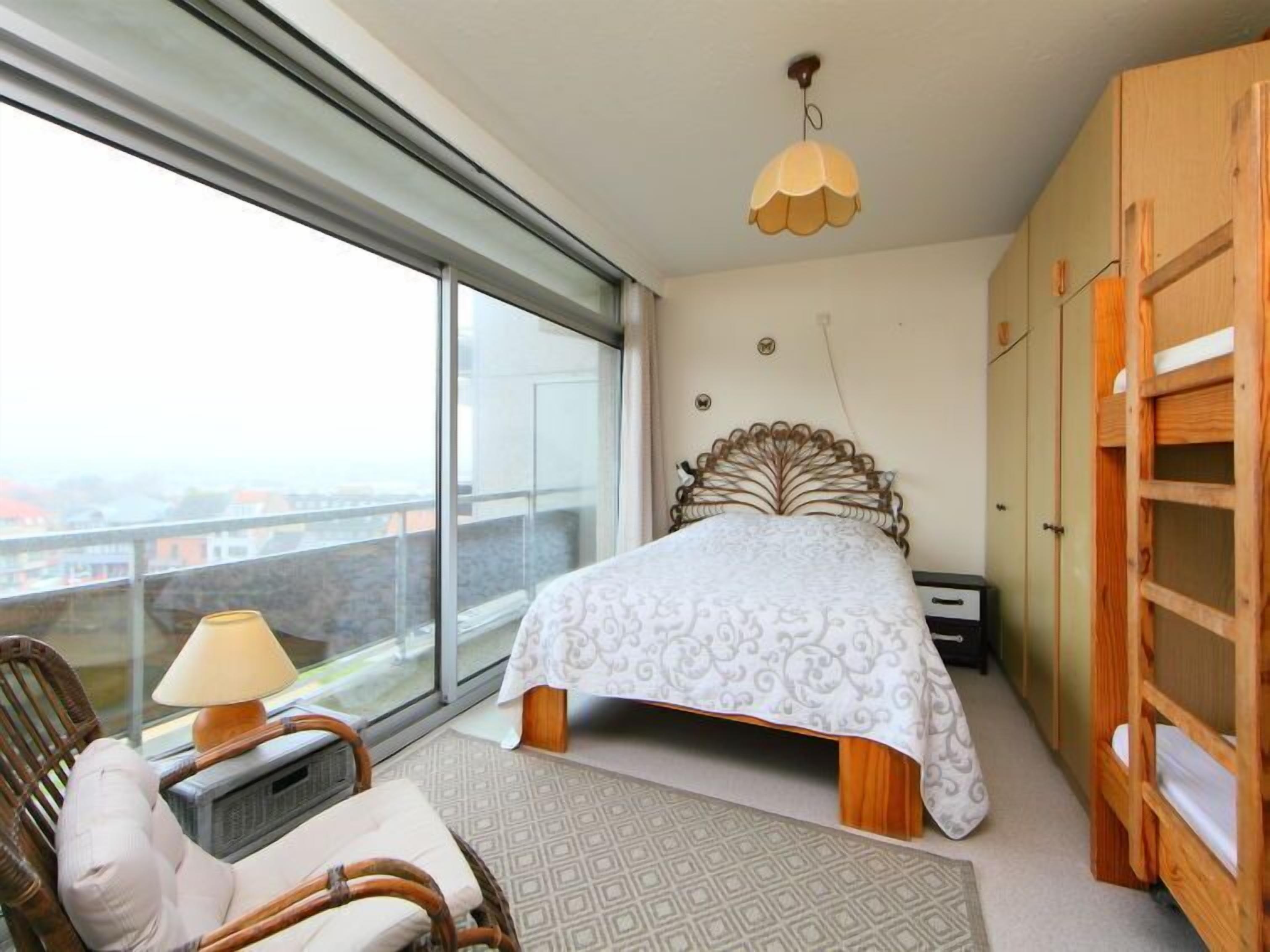 Une chambre avec une grande fenêtre, un lit recouvert d’une couvre-lit à motifs, une table de chevet avec une lampe, une armoire en bois et une vue sur les bâtiments à l’extérieur.
