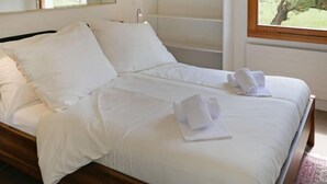 A neatly made bed with white linens and towels, a wooden headboard, and a window with a view of trees.