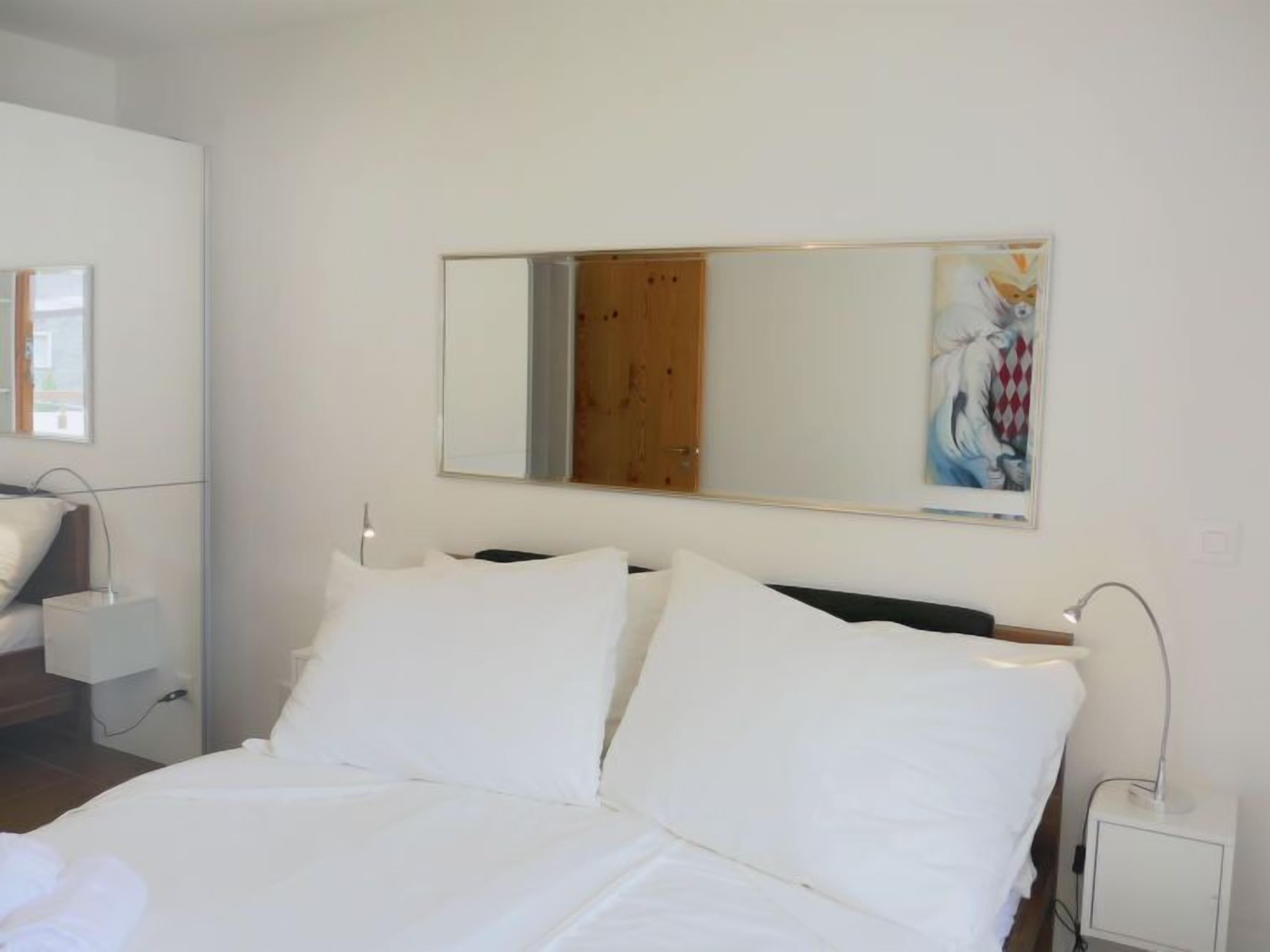 Une chambre moderne avec un grand lit, une porte en bois, une table de chevet blanche et un miroir fixé au mur.