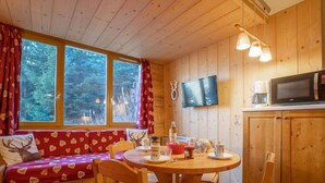 Ein gemütliches Blockhaus-Interieur mit einem Holzesstisch, Stühlen, einer Couch mit Kissen mit Herzmuster, einem Fernseher und einer Küchenzeile inklusive Mikrowelle.
