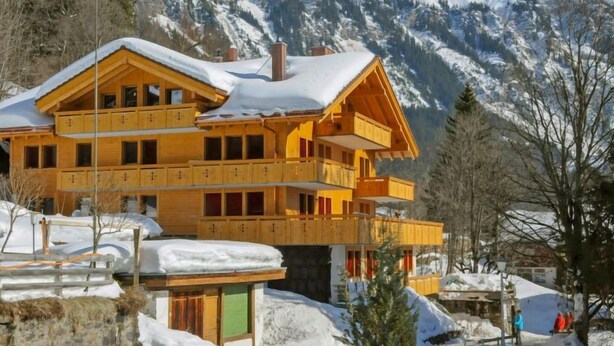 A multi-story wooden chalet with balconies, set against a snowy mountain backdrop.