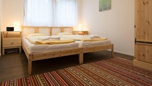 A bedroom with a wooden bed, two bedside tables with lamps, a window with sheer curtains, a wooden wardrobe, and a colorful striped rug.
