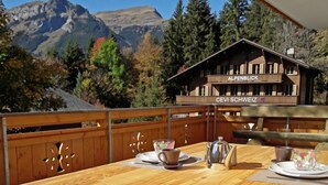 A wooden table with a view of mountains, a wooden building labeled "ALPENBLICK" and "CEVI SCHWEIZ", and a wooden fence.