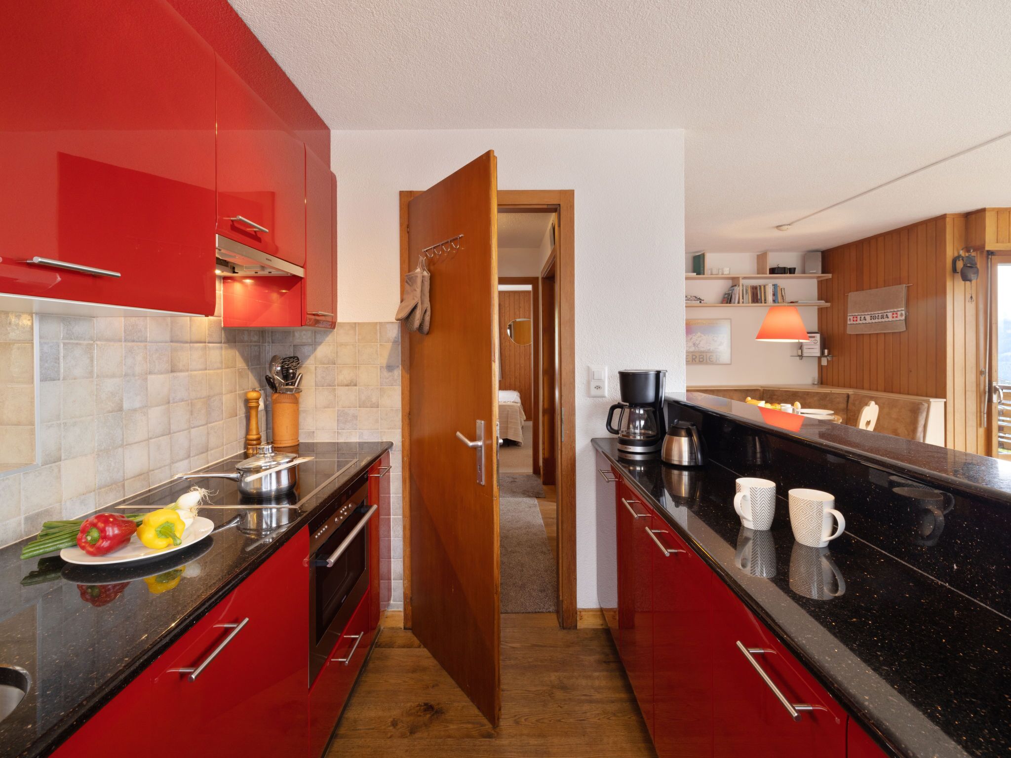 A modern kitchen with red cabinets, a black countertop, and a wooden door leading to a dining area.