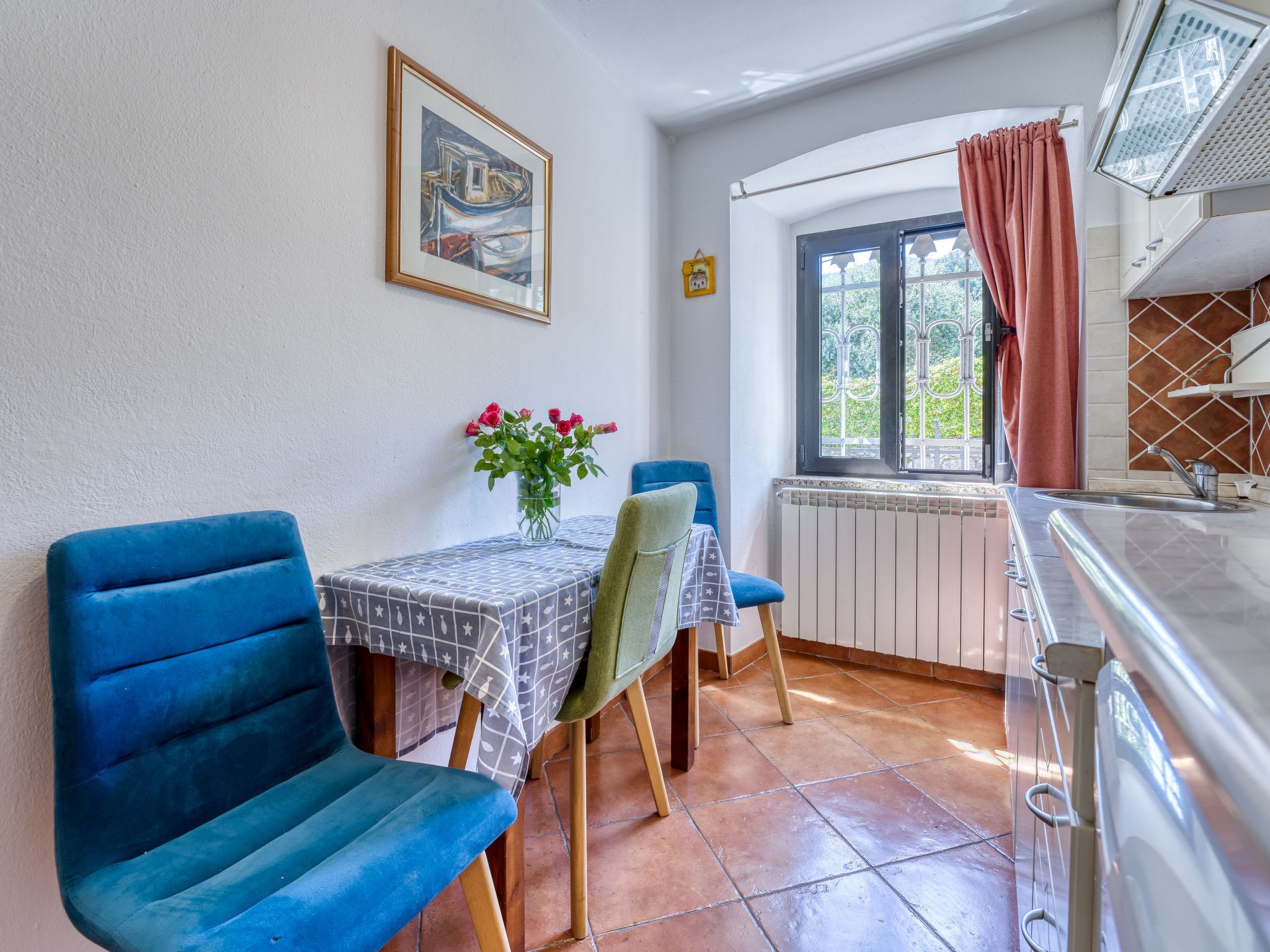 A dining area with blue chairs, a table with a patterned tablecloth, a vase with flowers, and a window with a view of greenery.