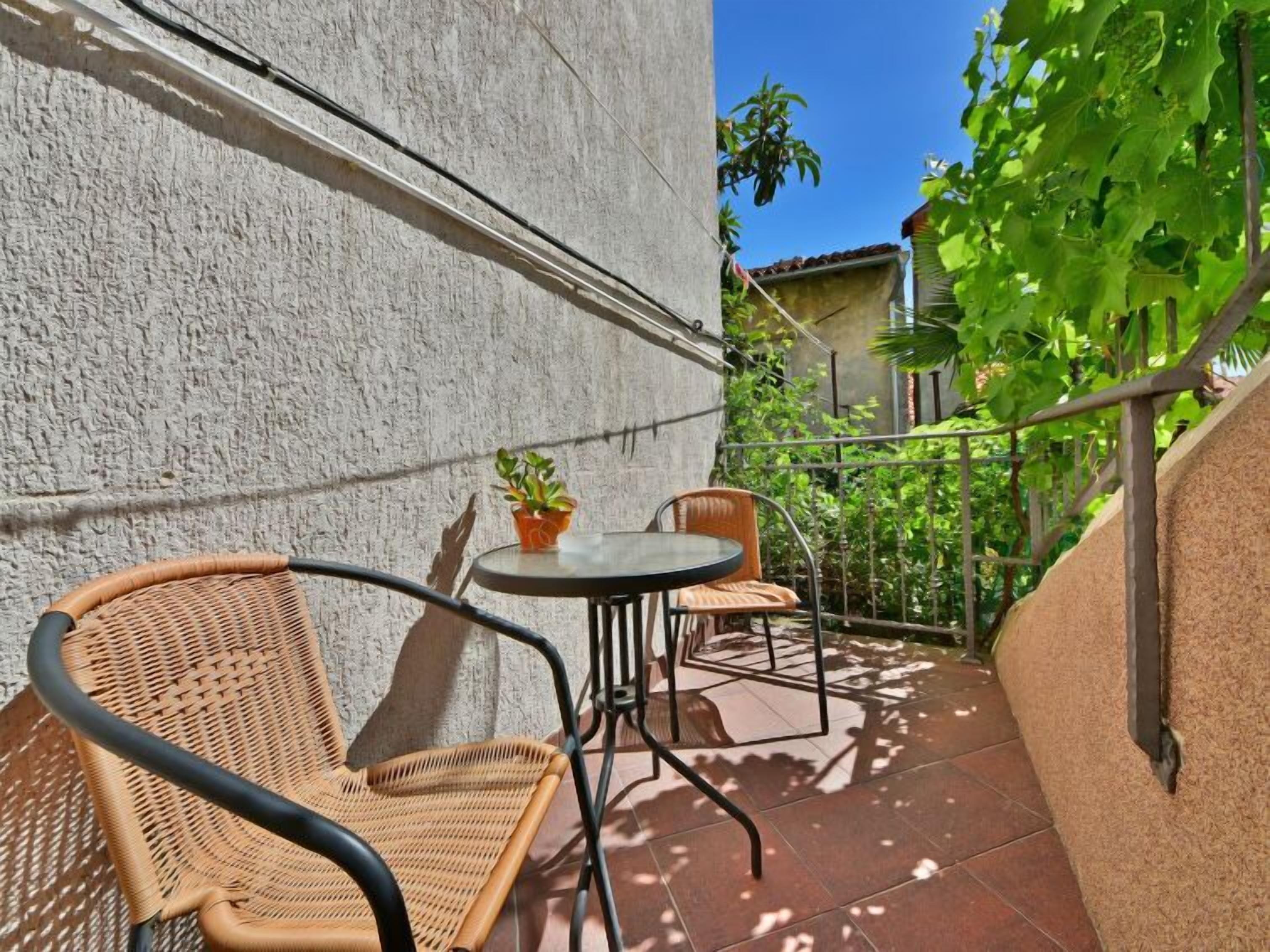 A small outdoor patio with a table and two wicker chairs, a potted plant, and a view of a garden with trees and a building in the background.
