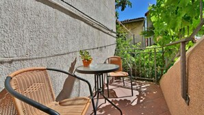 A small outdoor patio with a table and two wicker chairs, a potted plant, and a view of a garden with trees and a building in the background. - Lovrano by Interhome (Lovran)