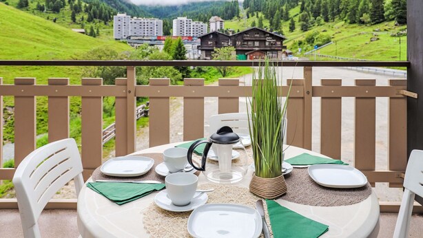 A balcony with a table set for two, overlooking a mountainous landscape with buildings in the distance.