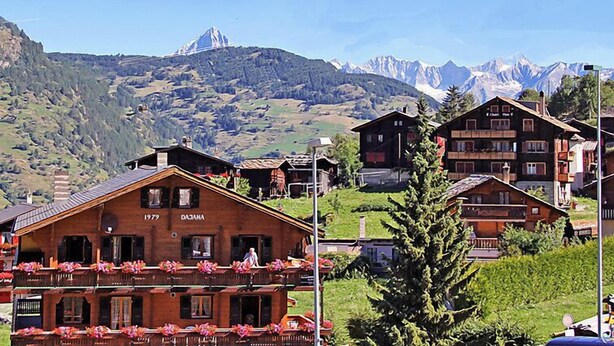 Ein traditionelles Holzhäuschen mit Balkon, geschmückt mit Blumen, vor einer Berglandschaft und blauem Himmel.