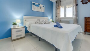 A bedroom with a bed, a nightstand, a lamp, a potted plant, and a window with curtains. - El Santiago II by Interhome (Roses)