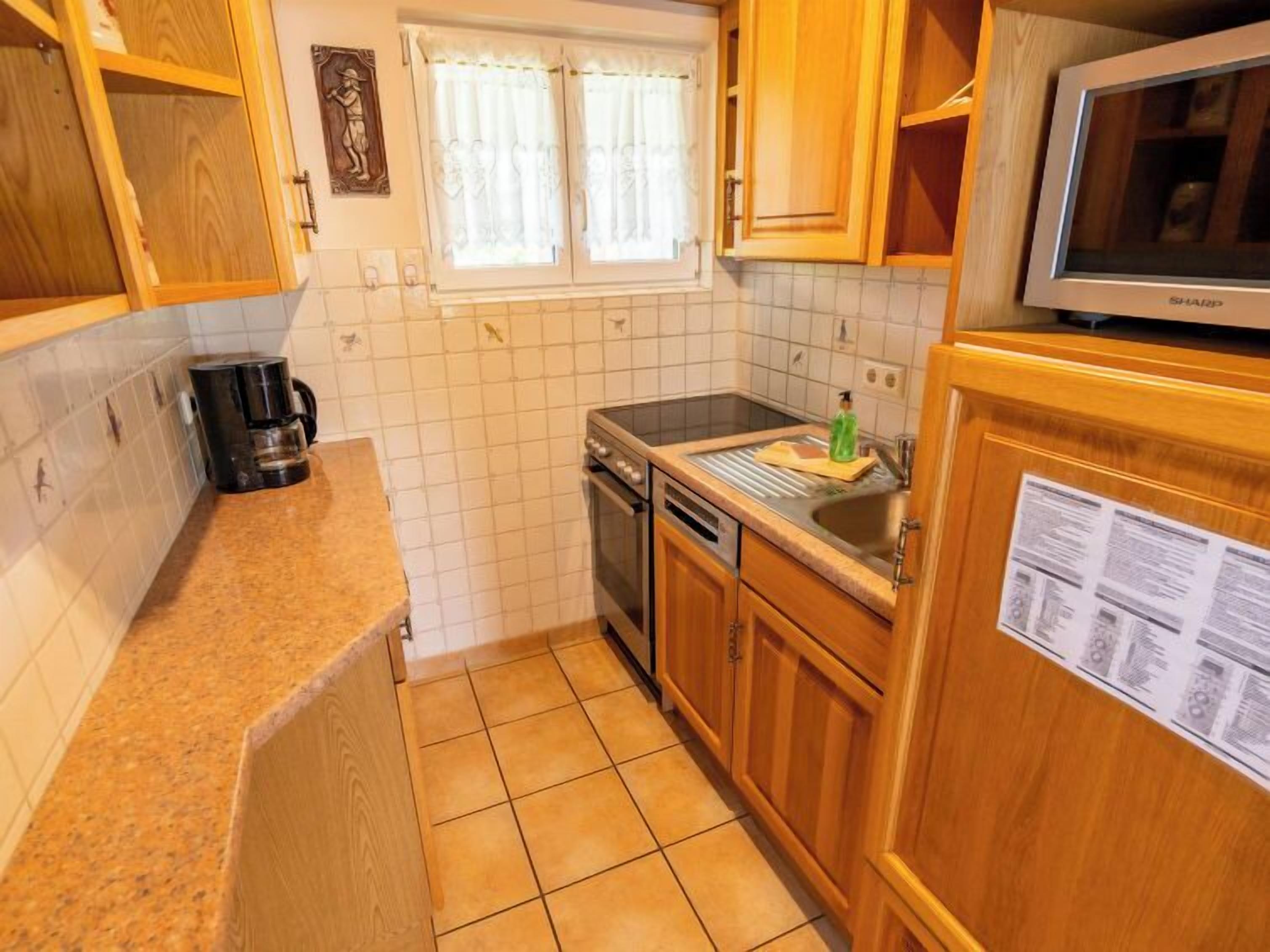 A compact kitchen with wooden cabinets, a built-in oven, a coffee maker, and a window with curtains.