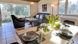 A dining area with a table set for four, a sofa, and a view of trees through large windows. - Wanderlust by Interhome (Dittishausen)