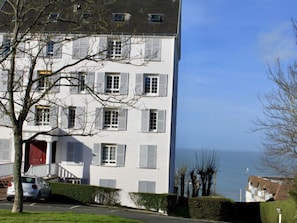 A multi-story building with white walls and shutters, a red door, and a view of the sea.