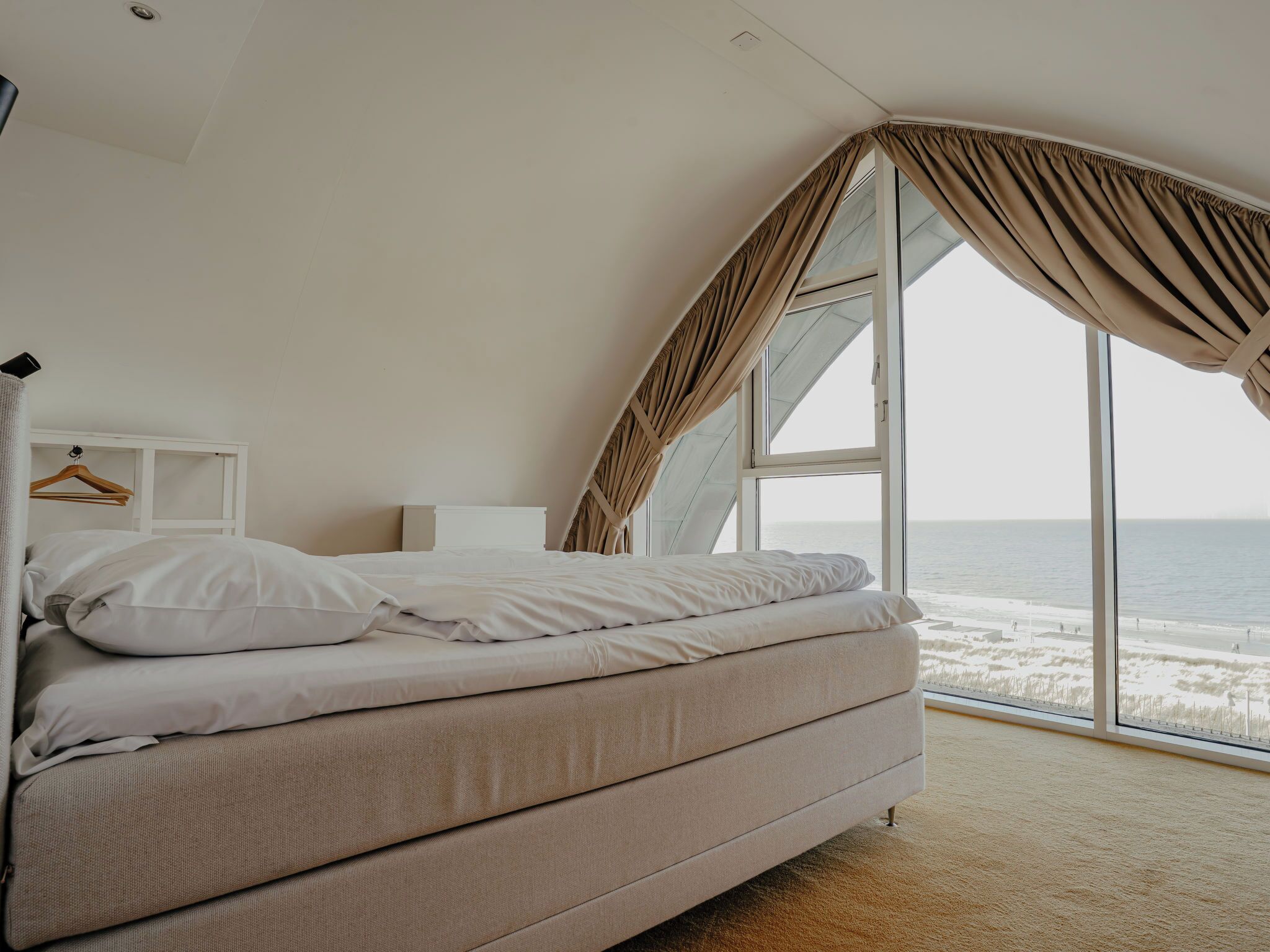 A bedroom with a bed, a nightstand, a hanging rack, and a large window with curtains.