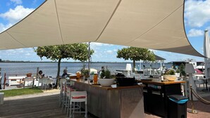 An outdoor bar area with a wooden deck, white bar stools, and a large canopy providing shade. - Dorregeest by Interhome (Uitgeest)