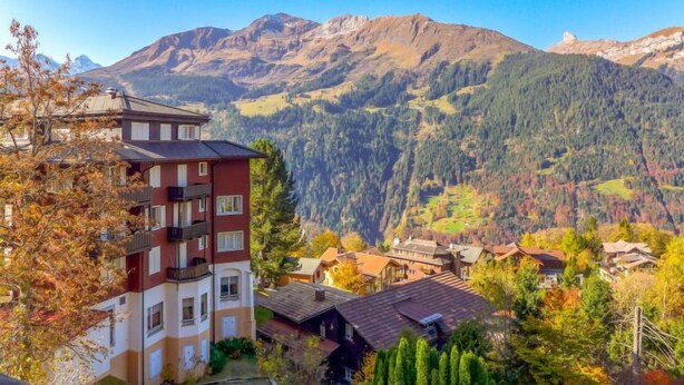 A multi-story building with balconies, surrounded by autumn-colored trees and a mountainous landscape. - Apartment Biwak by Interhome (Wengen)