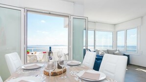 A modern dining area with a wooden table set for four, large windows offering a view of the sea, and a balcony with a blue sofa.