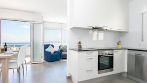 A modern kitchen with white cabinets, a built-in oven, and a dishwasher. There is a dining area with a table and chairs, and a blue chair with a white cushion.