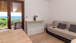 Une pièce avec une table en bois, un canapé blanc avec des coussins, une petite armoire blanche et une vue sur l’océan par une porte coulissante.