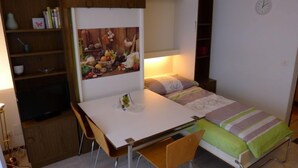 A compact room with a bed, a desk, and a chair. There is a shelf with books and a lamp.
