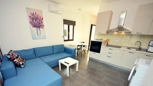 A modern living room with a blue sofa, a small white table, and a kitchen area with white cabinets and a stove. - Molo Lozna by Interhome (Brač/Postira)