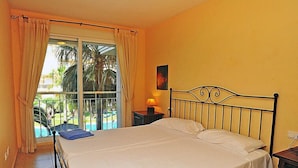 A hotel room with a bed, a nightstand, a lamp, and a view of a pool and palm trees through a window. - Nou Fontana by Interhome (Jávea)
