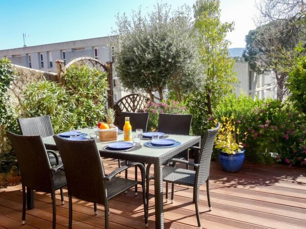 An outdoor dining area with wicker chairs and a table set for two, surrounded by greenery and a building in the background.